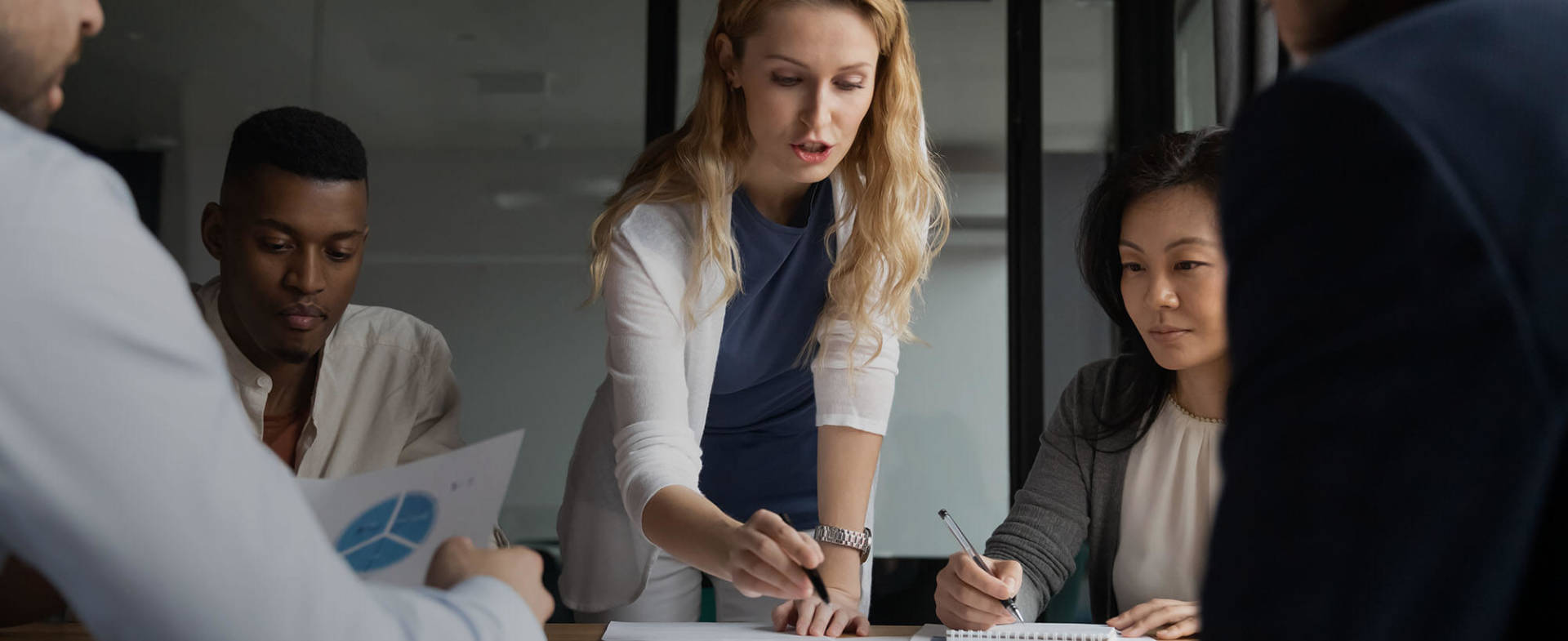 Marketing manager leading a team meeting and pointing a pen at a sheet of paper while four other coworkers look on.