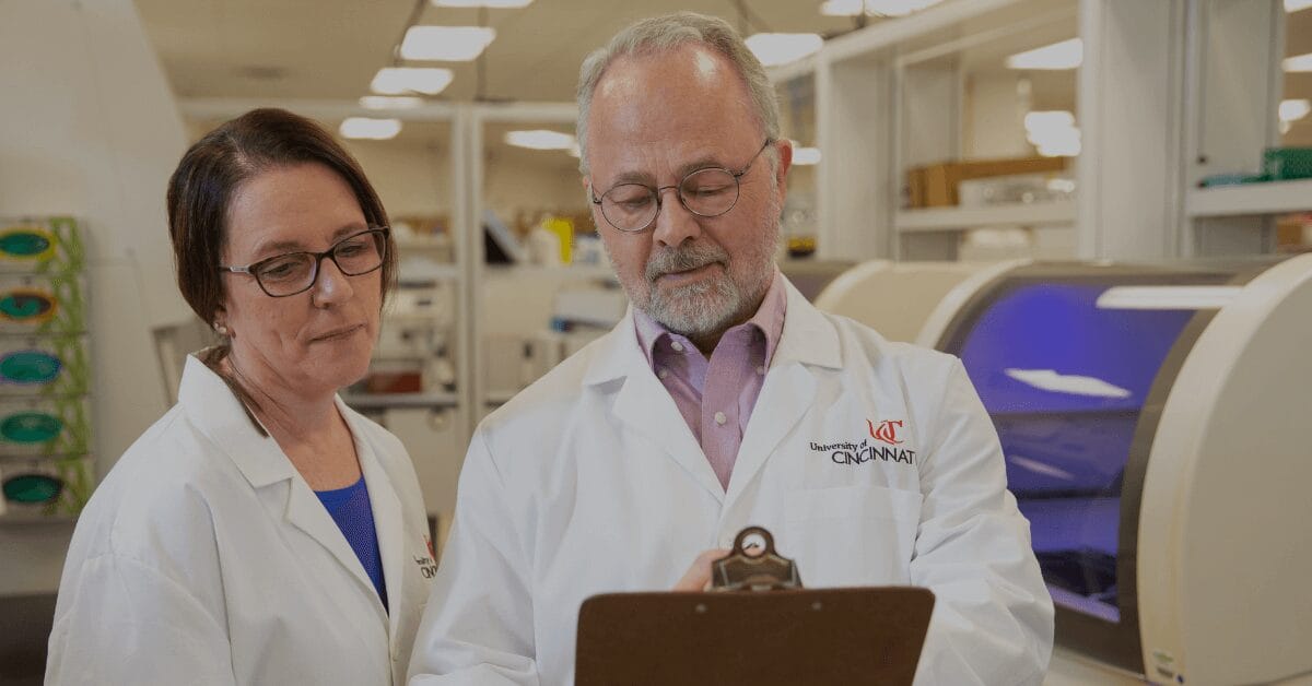 Two medical laboratory science professionals wearing University of Cincinnati lab coats discussing test results in a modern clinical laboratory.