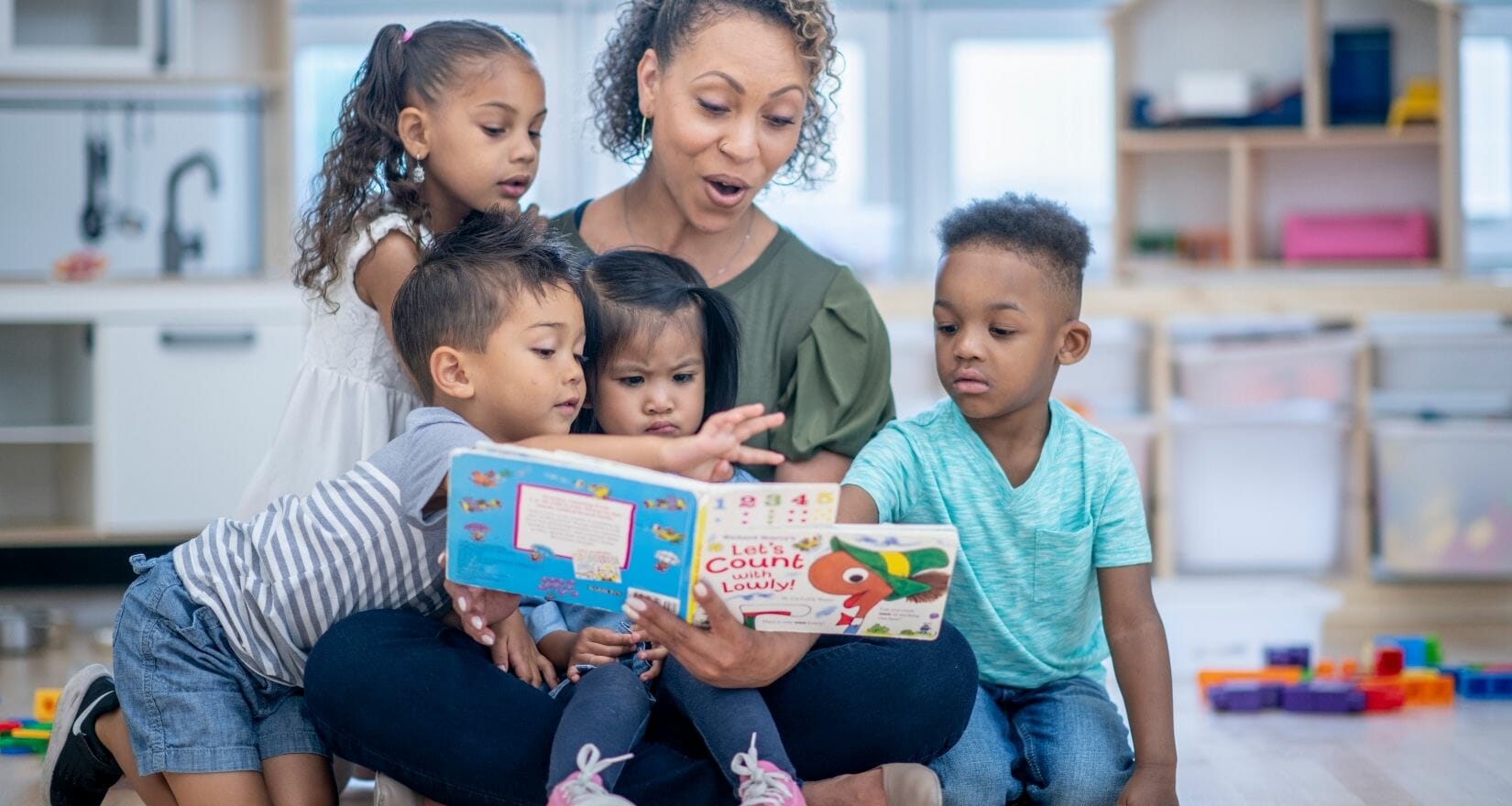 A woman reads a book to a group of young children who are sitting on her lap and around her.