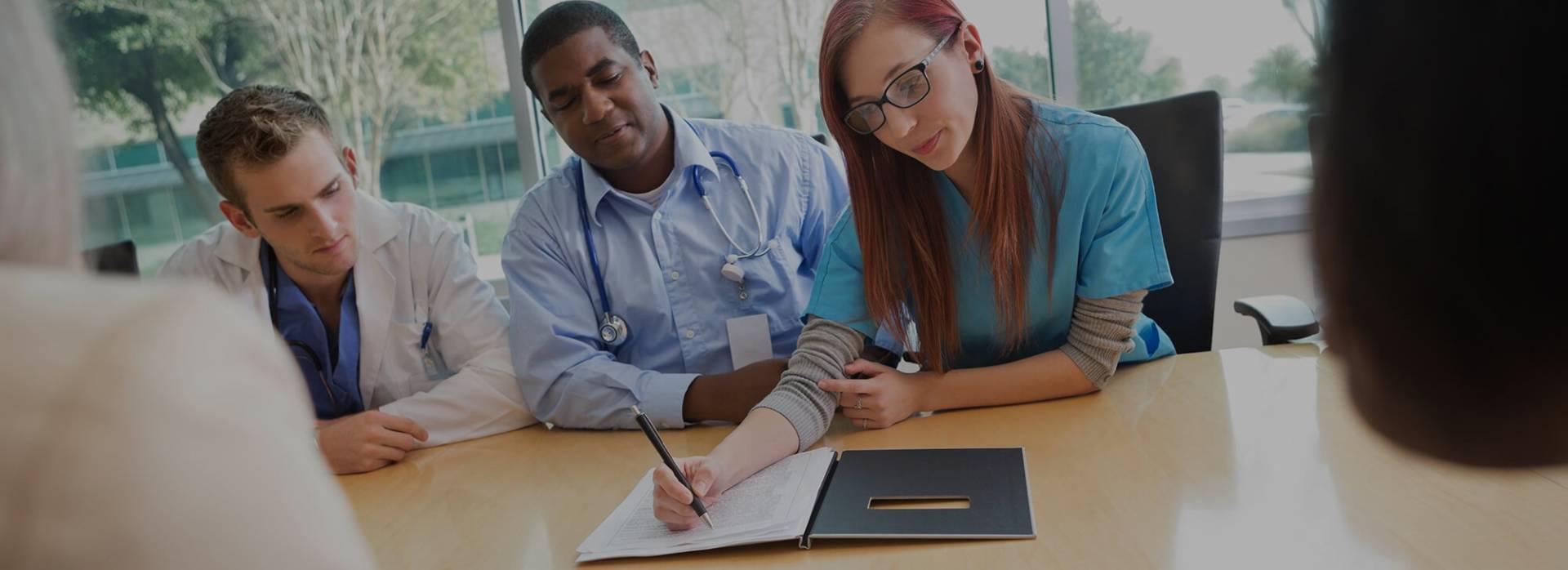 Three health professionals are sitting at a wooden table while one of them writes in a notebook.