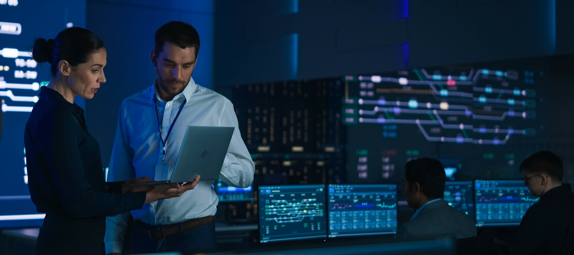 Two cybersecurity professionals stand and look at an open laptop screen while other people are working on dual-monitor computers in the background.
