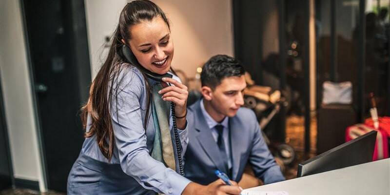 Hospitality management professional answering the phone while another observes a computer screen.