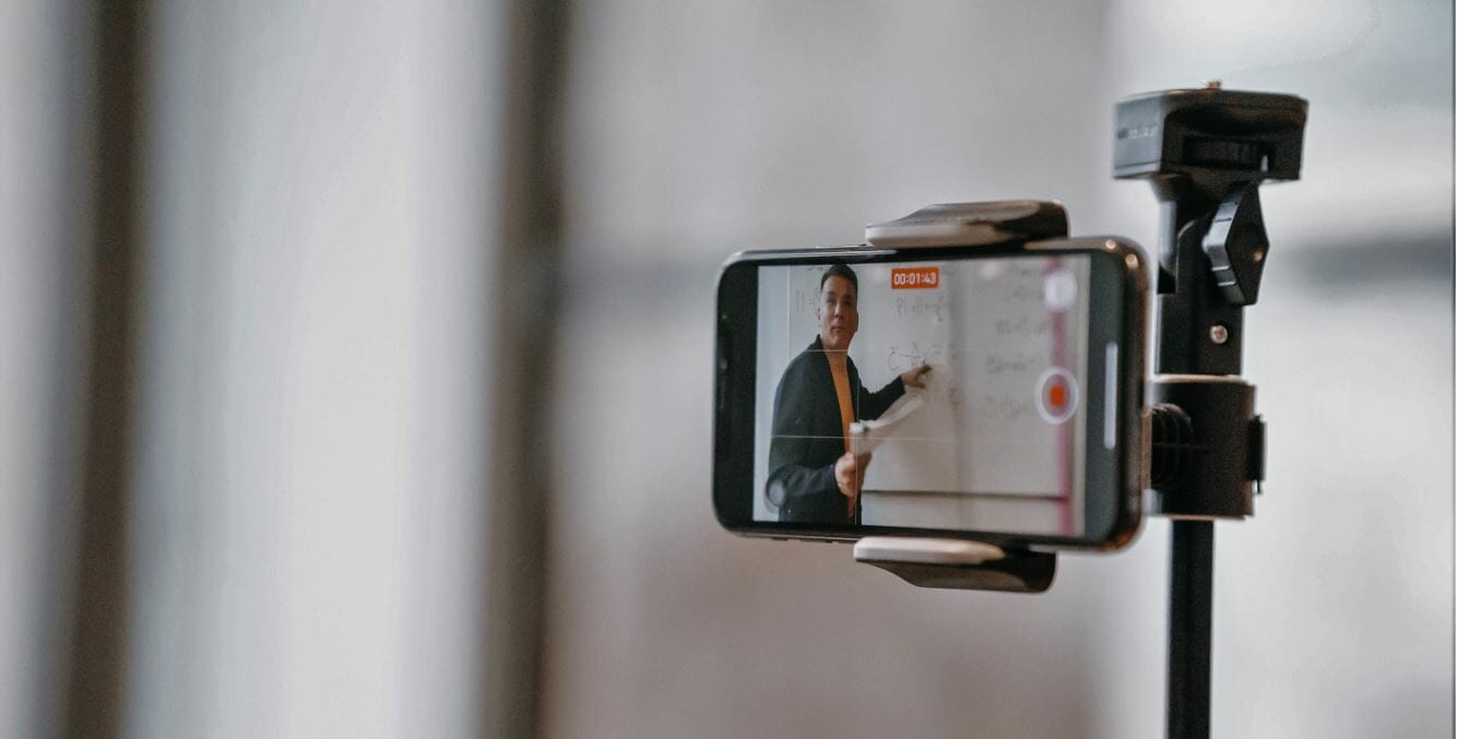 Educator recording an online lesson using a smartphone camera mounted on a tripod, demonstrating concepts on a whiteboard.