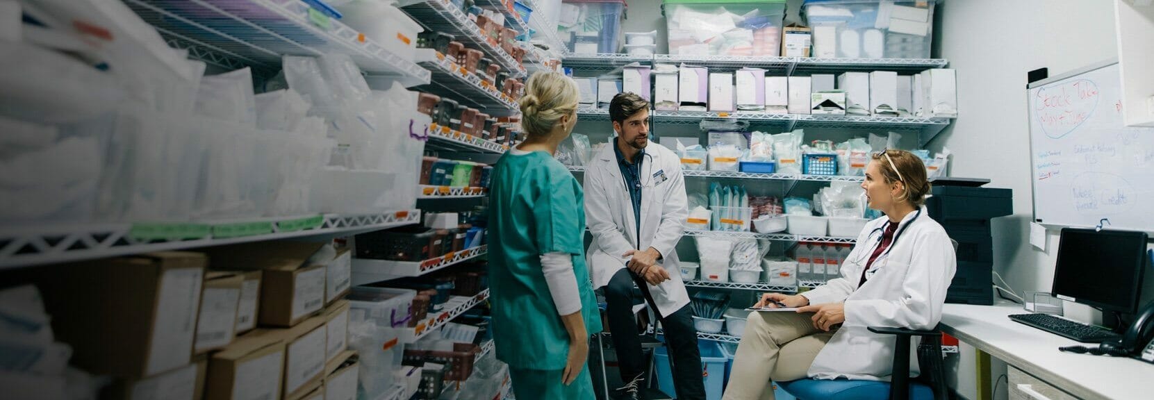 Pharmacy professionals in lab coats and scrubs collaborating inside a medical supply room, discussing inventory and management strategies, representing leadership and organization in pharmacy practice.