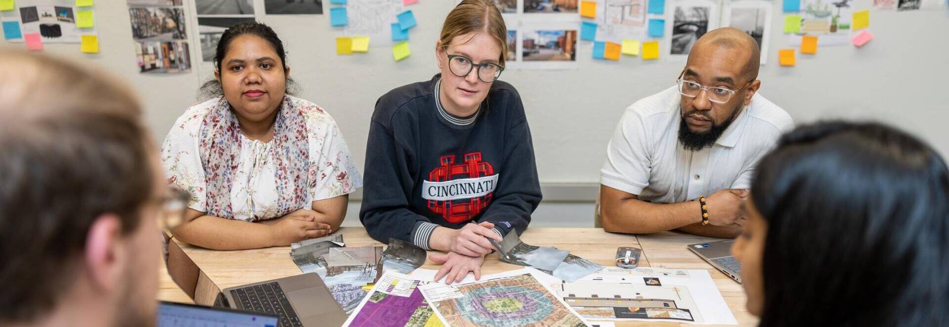 Graduate students collaborating on a city planning project at a table covered with maps and design layouts, with colorful notes and urban development plans on the wall behind them, representing teamwork in the Master of City Planning program.