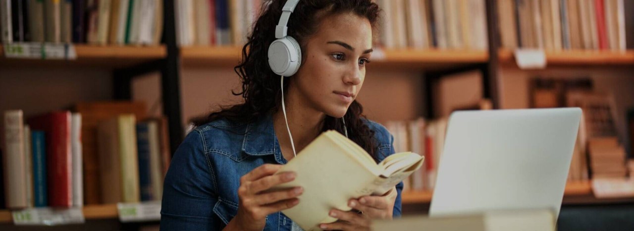 A English as Second language teacher wearing headphones and reviewing teaching materials on a laptop with a book in hand.