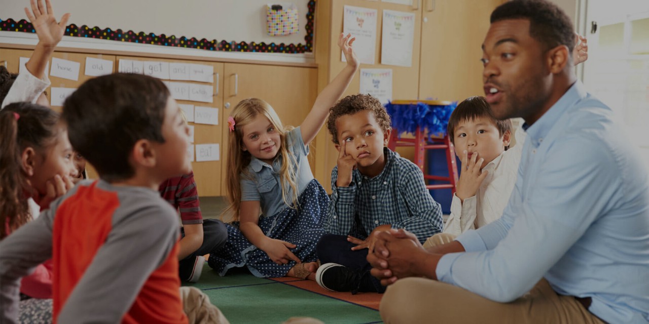 A teacher and a group of young students are all sitting on the floor each with their legs crossed while the teacher speaks to the group.