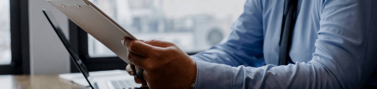 This close-up image shows the torso and arms of a person wearing a blue-collared shirt with a tie while holding a clipboard next to an open laptop.