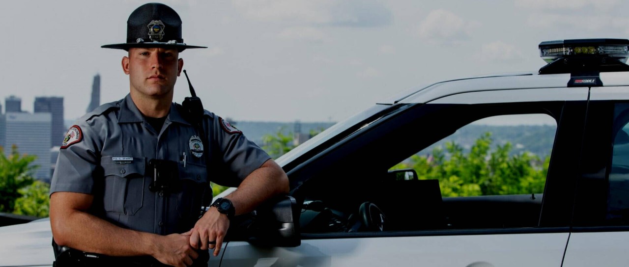 A police officer in a hat poses next to his patrol car while leaning on the driver's side mirror with his left forearm.